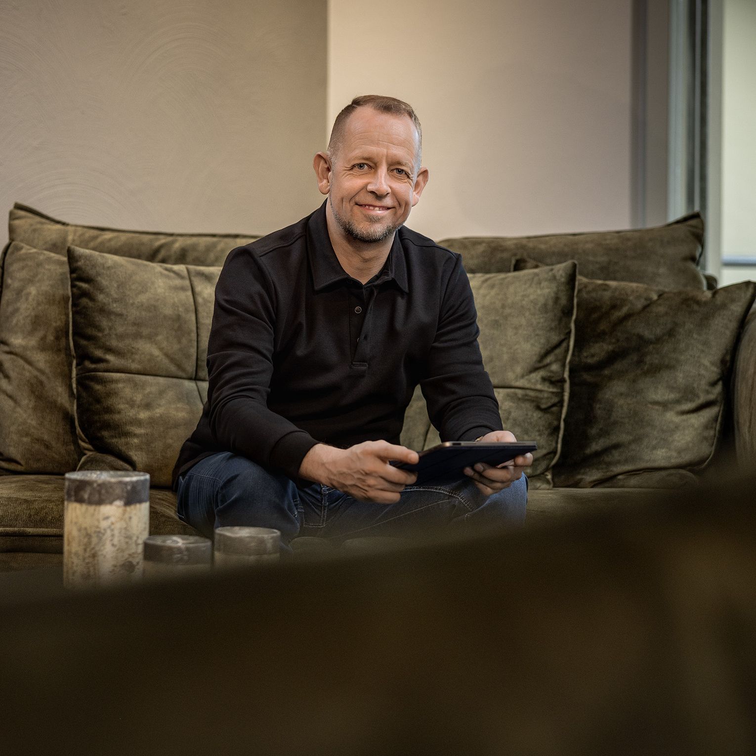 Joachim Köllmann sitzt lächelnd auf einem Sofa mit einem iPad in der Hand.